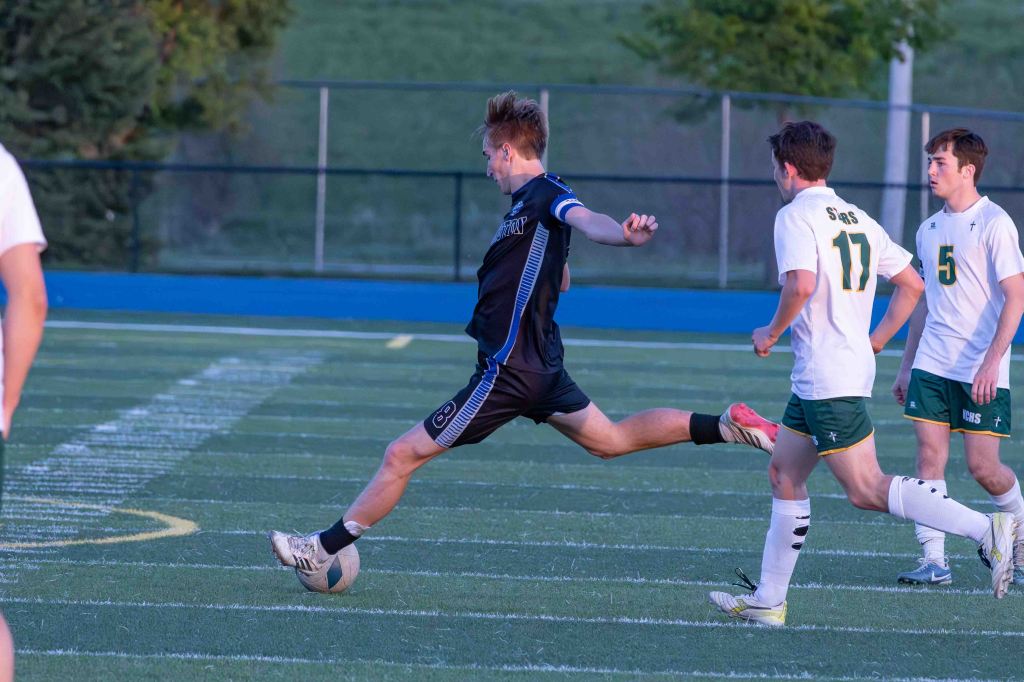 A soccer player in a black uniform kicks a soccer ball on a field, with other players in action nearby.