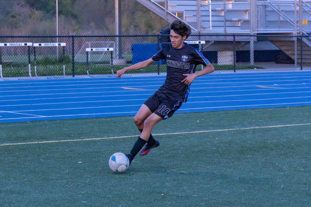 A young male soccer player in a black uniform dribbling a soccer ball on a field with blue track markings.
