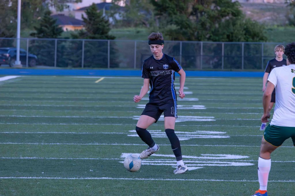 A soccer player in a black uniform dribbles a ball on the field during a match, with another player in a white uniform nearby.