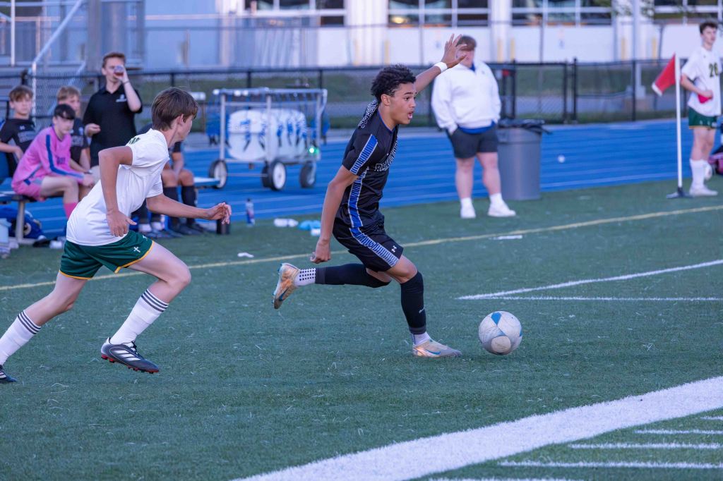 A young soccer player in a black jersey dribbles a ball on the field, pursued by an opponent in a white and green uniform during a game.