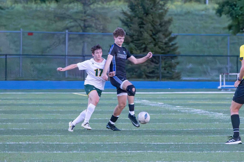 A soccer match in progress with two players competing for the ball. One player in a black uniform is about to kick the ball while another player, wearing a white jersey with the number 17, is attempting to challenge him. The scene is set on a green turf field.