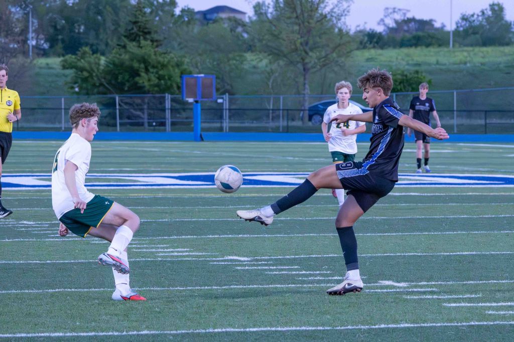 A soccer match in progress with two players competing for the ball. One player in a white jersey and green shorts is attempting a tackle, while the player in a black jersey is kicking the ball. A referee is visible in the background.