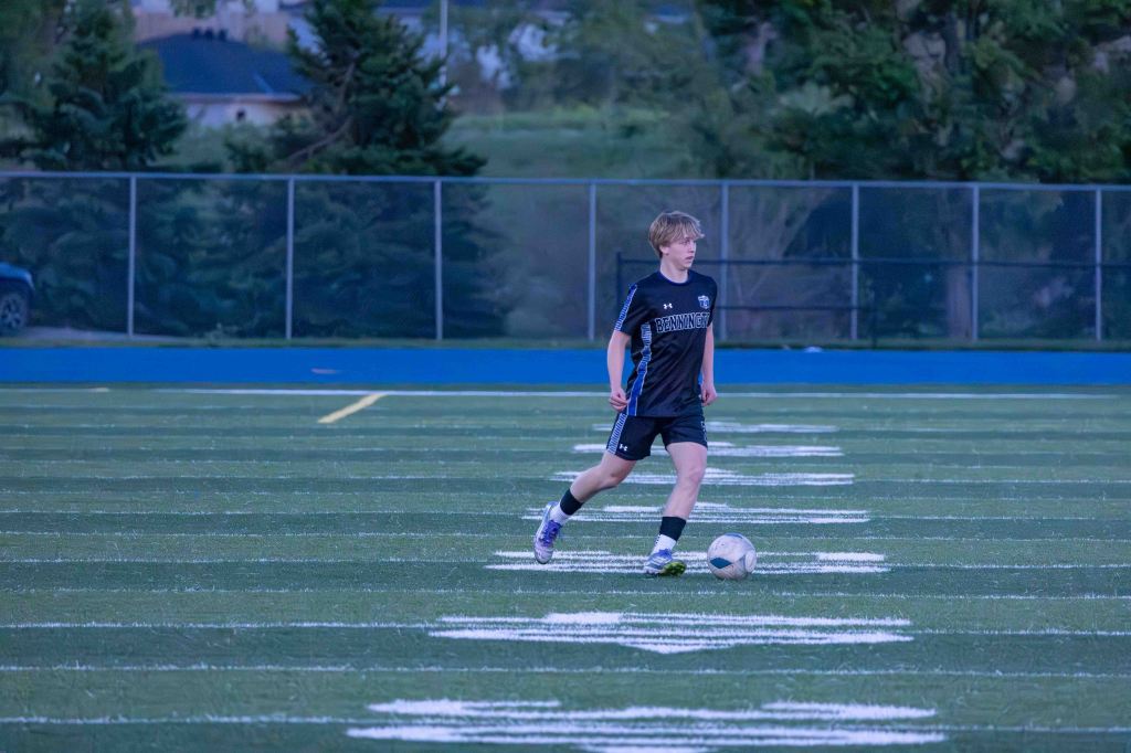 A young male soccer player wearing a black jersey and shorts running on a sports field with a soccer ball at his feet.