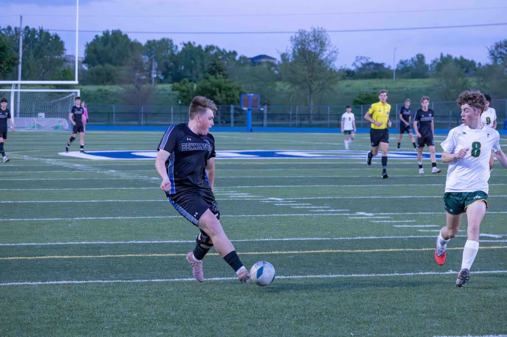 A soccer match in progress, featuring a player in black kicking the ball, while an opponent in white and green runs toward it. Team members are visible in the background, with goalposts and a green field.