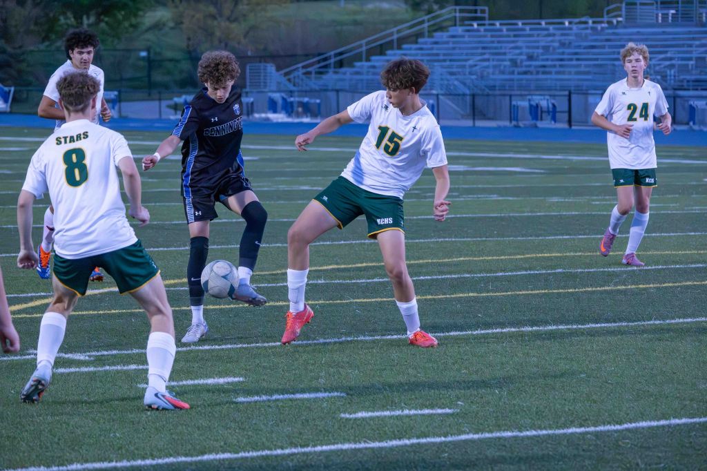 High school soccer match with players in action on the field. One player in black is trying to kick the ball while two players in white and green attempt to defend.