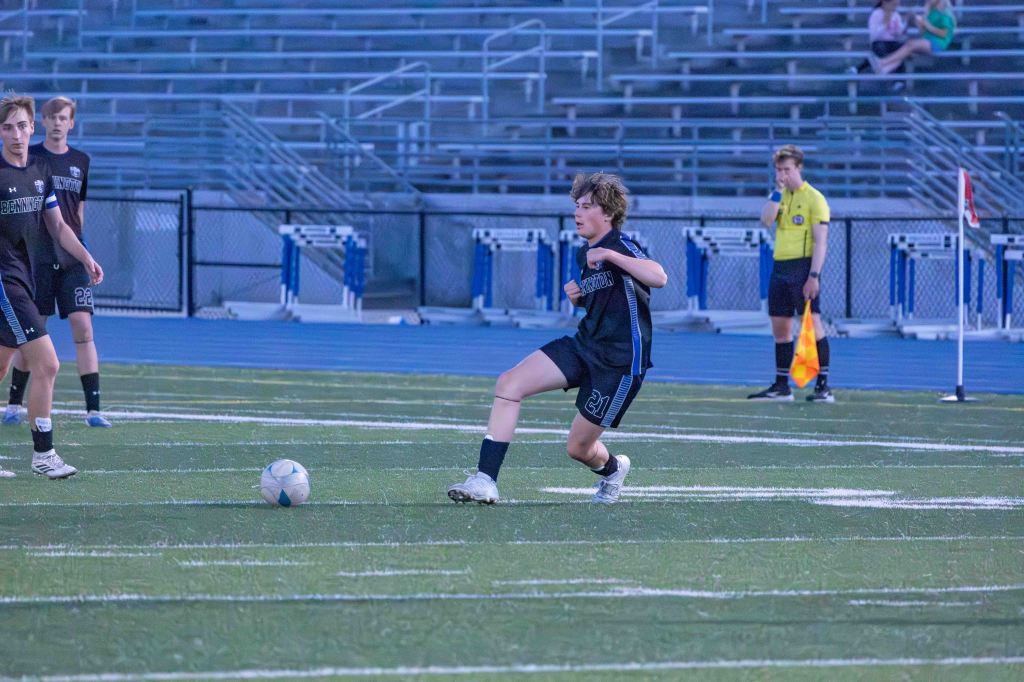 A soccer player in a blue uniform is kicking a soccer ball on a field, with teammates and an official in the background.