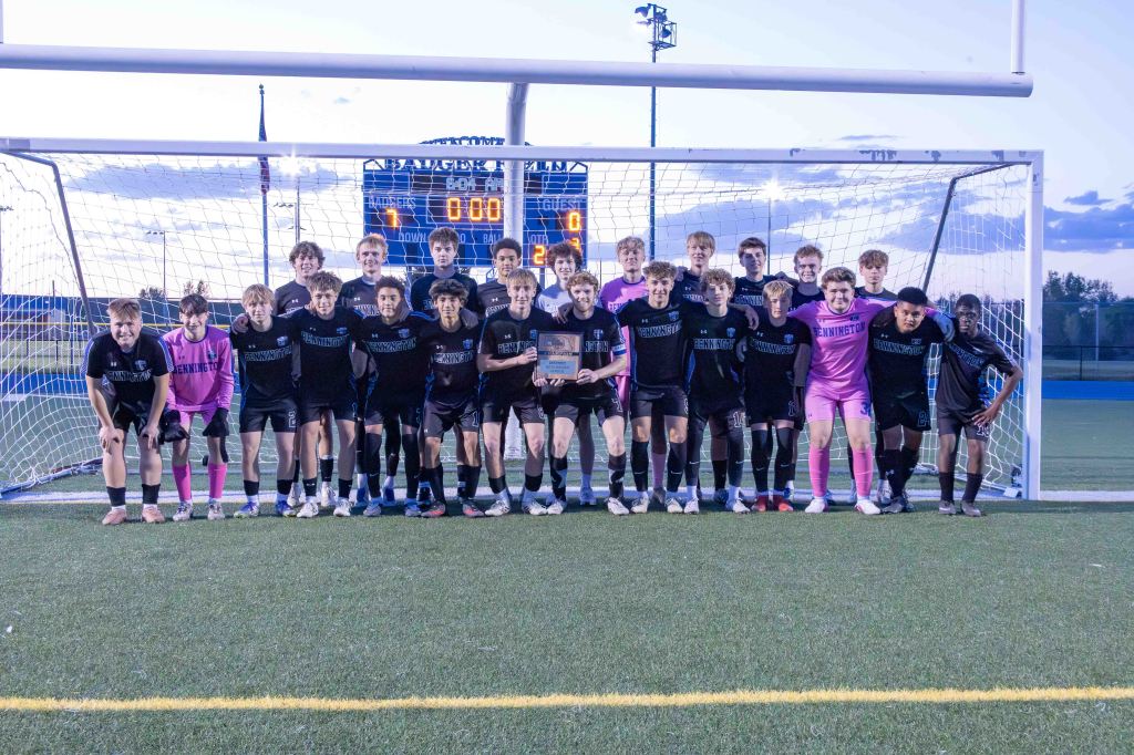 A group photo of a youth soccer team in black uniforms standing in front of a goalpost, with a scoreboard showing 0-0. The team includes players celebrating and a goalkeeper in pink, captured on a grassy field during dusk.