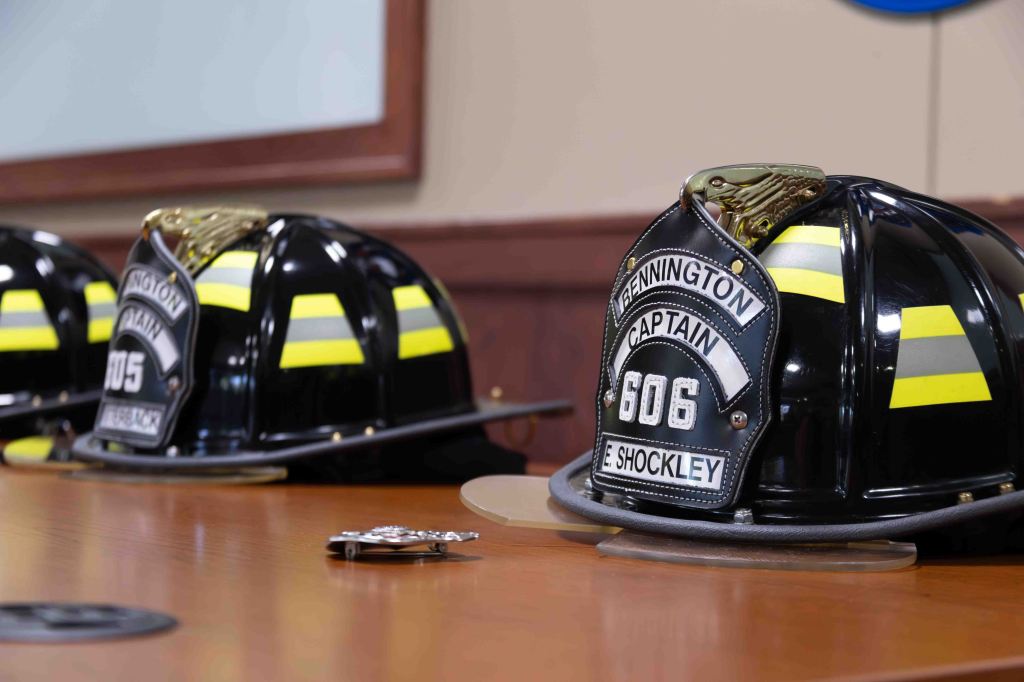 Three fire helmets with yellow stripes and badges on a wooden table, with a reflective surface and a blurred background.