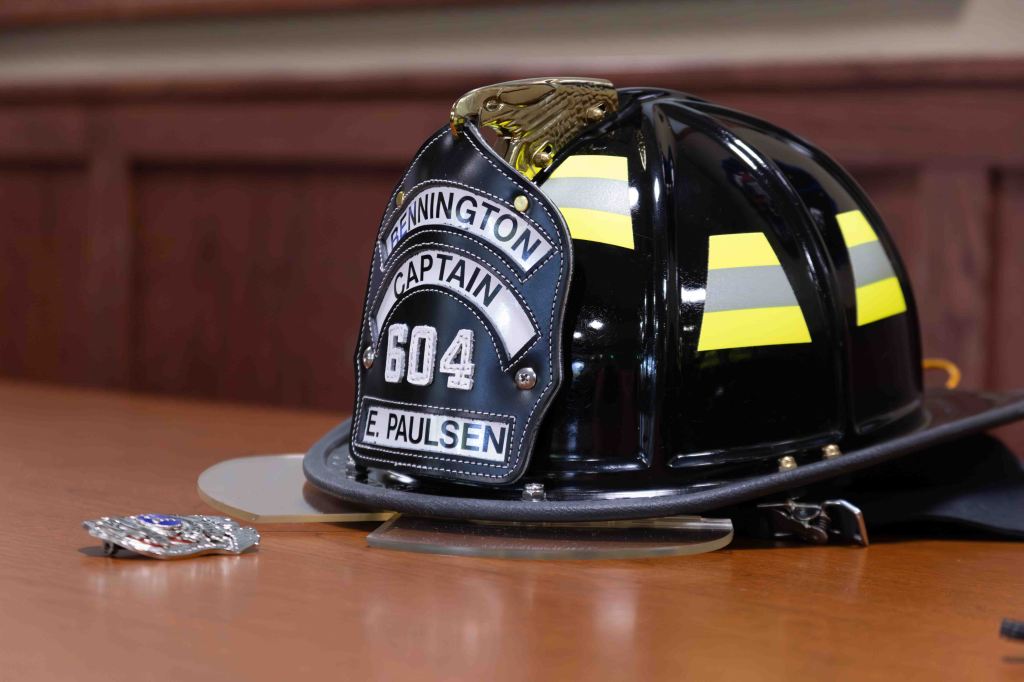 A firefighter's helmet with yellow reflective stripes, featuring a badge that reads 'Bennington CAPTAIN 604 E. Paulsen' and a silver police badge placed beside it on a wooden table.