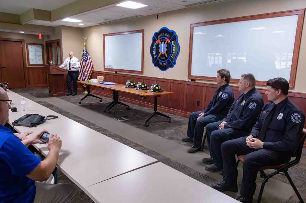 A fire chief speaks at a podium in a conference room with an American flag, while three firefighters in uniforms sit nearby. A table displays helmets and materials, with a wall-mounted emblem in the background.