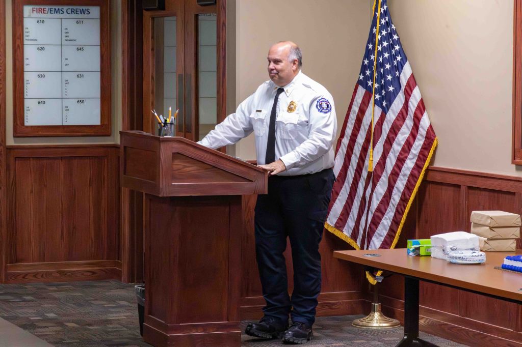 A man in a police uniform stands at a podium in a room with a wooden interior, next to an American flag. A table nearby has wrapped gifts and supplies.