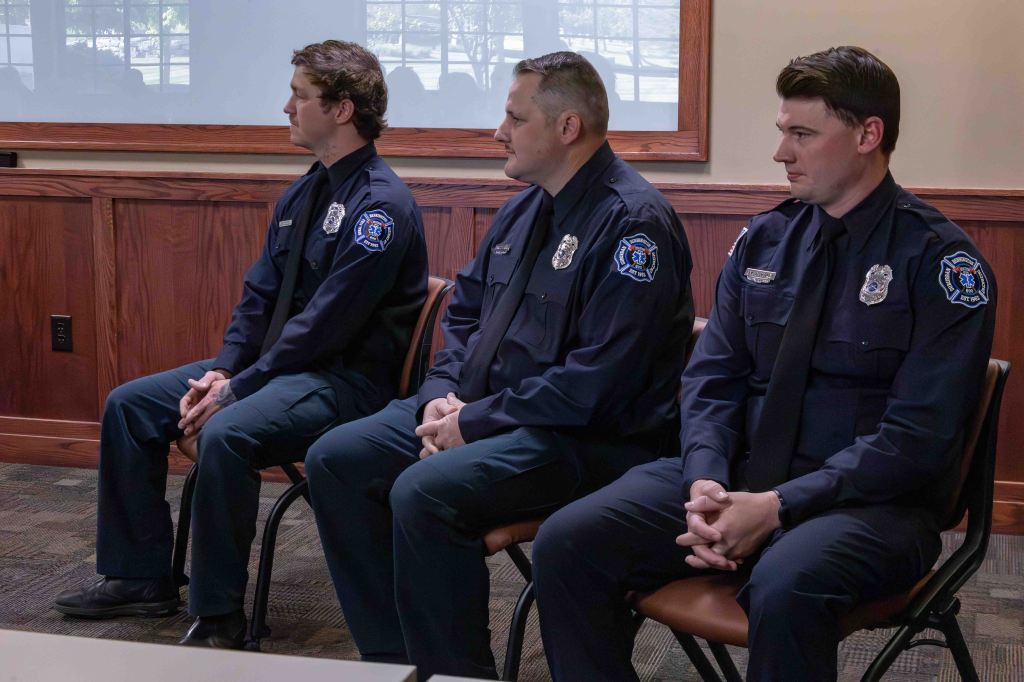 Three police officers sitting in a row, wearing uniforms, in an indoor setting with wooden paneling and large windows.