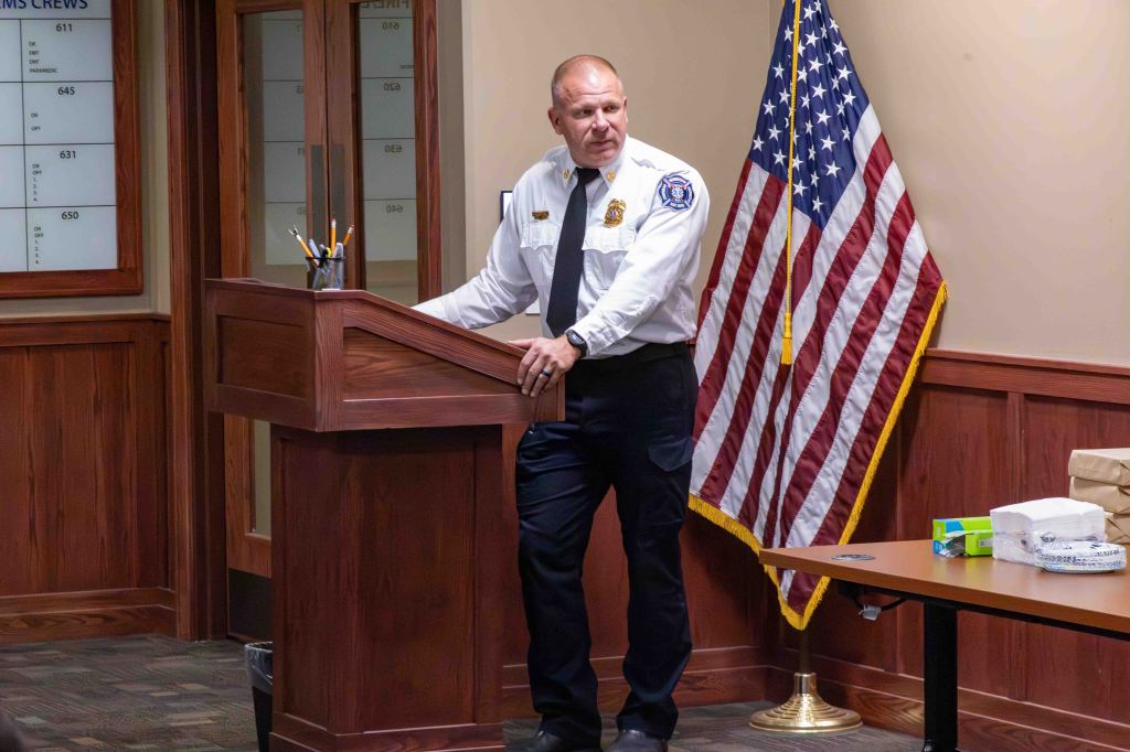 A uniformed officer standing at a podium in an office setting, with a U.S. flag in the background.