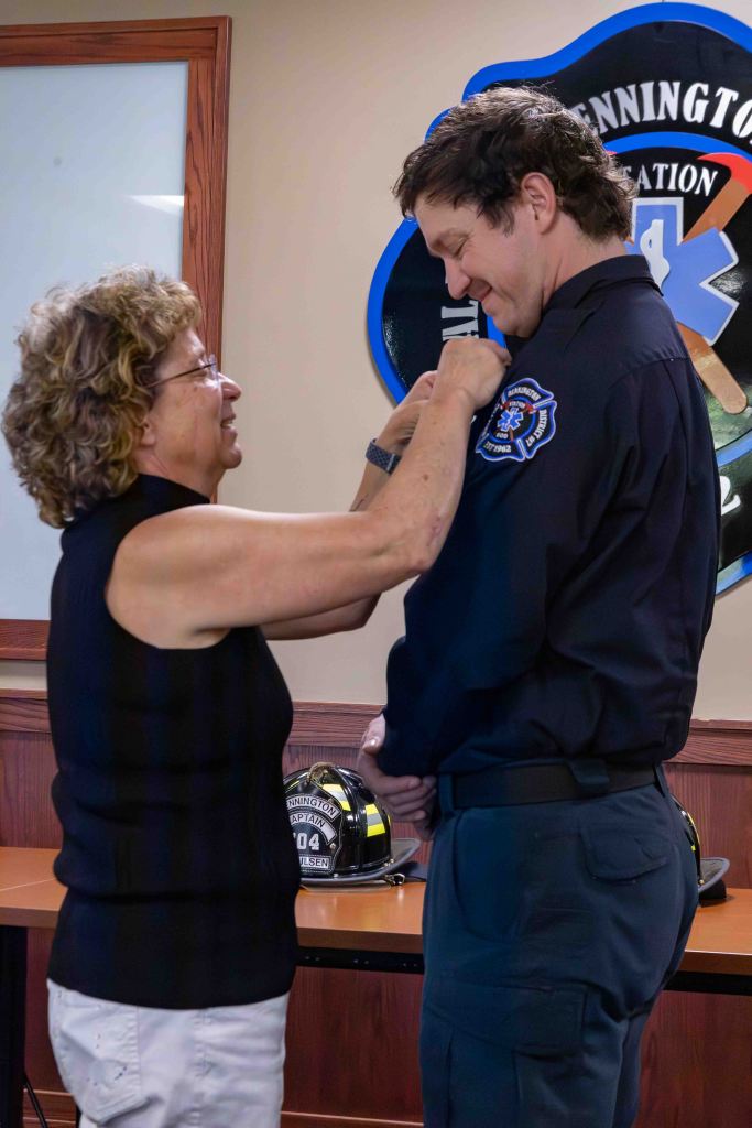 A woman pins a badge onto a man's shirt during a ceremony, with a fire helmet visible in the background.