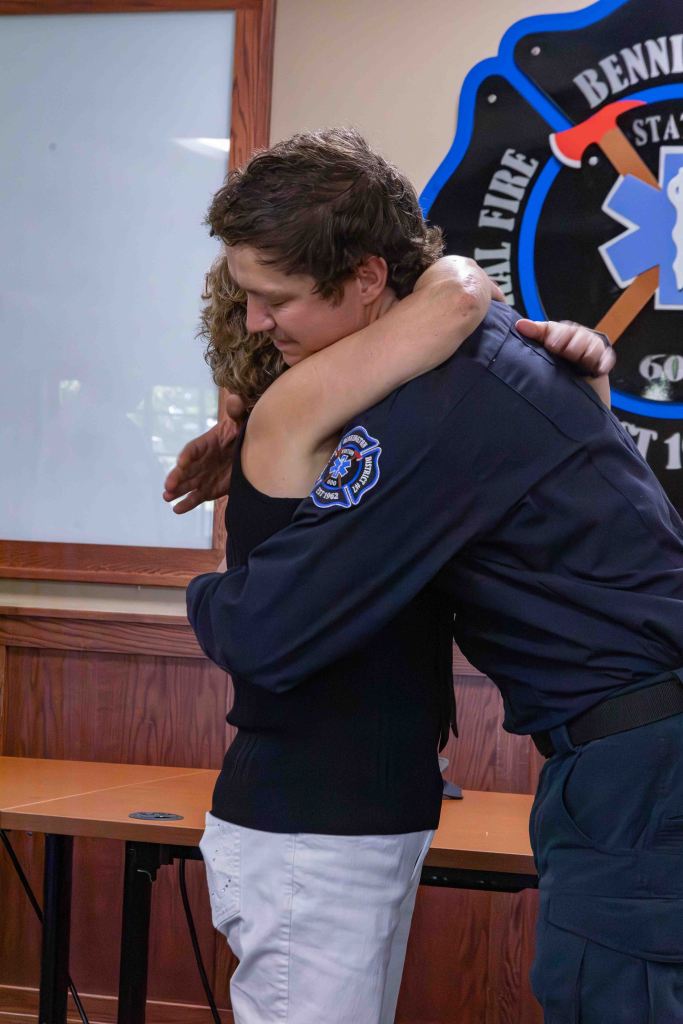 A heartfelt hug between a woman in a black top and a man in a fire department uniform, set in a room with wooden accents and a fire department emblem in the background.