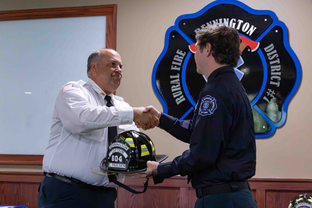 A fire captain in uniform shakes hands with a fire chief while receiving a firefighter helmet during a ceremonial event, with a district emblem displayed in the background.