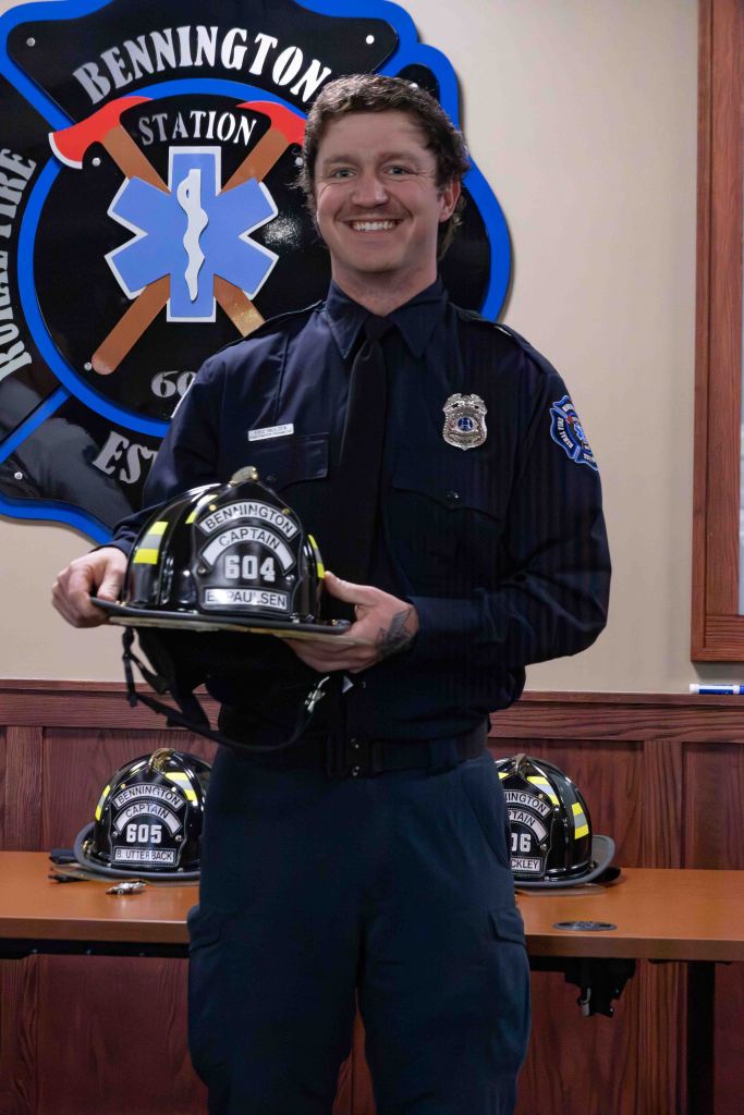 A firefighter in uniform smiles while holding a helmet with the number 604. Behind him is a badge displaying the Bennington Fire Station emblem and several additional helmets on a table.