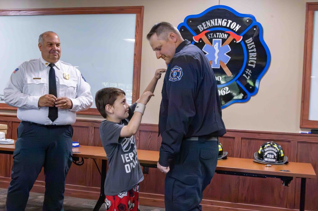 A young boy wearing a gray shirt and red shorts is pinning a badge on a firefighter in a black uniform, while a man in a white shirt and black tie watches with a smile. The background features a logo for the Bennington District and a table with firefighter helmets.