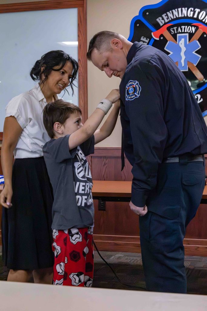 A young boy in a t-shirt and patterned shorts is pinning a badge on a firefighter's uniform, while a woman smiles at them in a meeting room decorated with a fire station emblem.