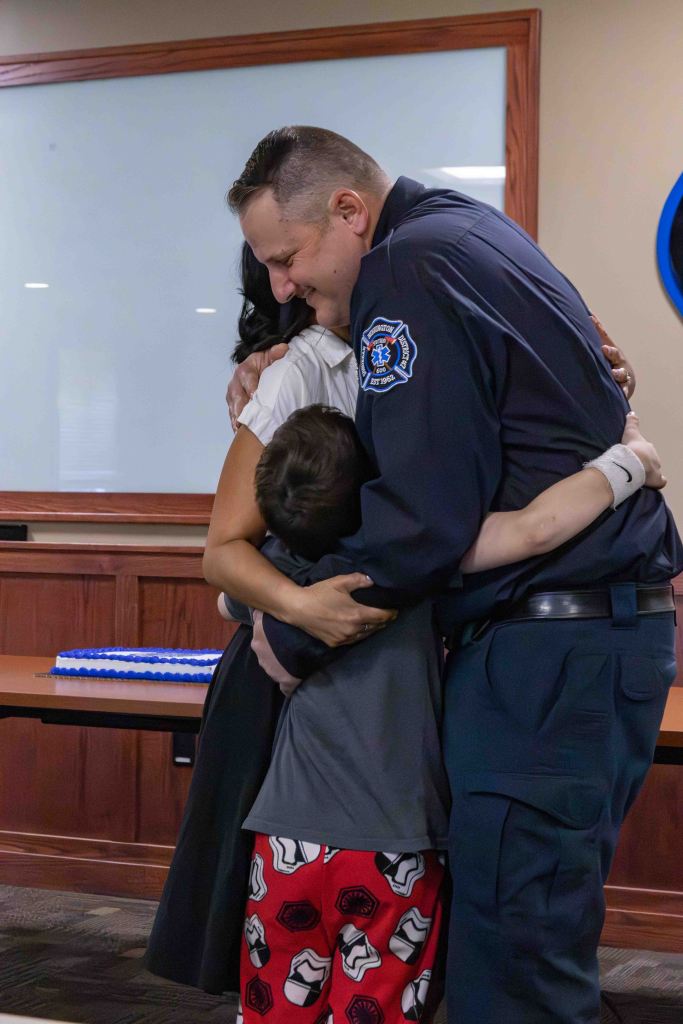 A firefighter embraces a woman and a child in a warm, emotional scene, celebrating a special moment together.