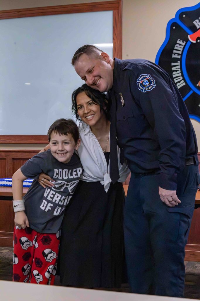 A cheerful firefighter poses with a woman and a young boy in a casual setting, all smiling together. The boy is wearing a 'Cooler Version of' t-shirt and pajama pants, while the woman is dressed in a black dress. The firefighter is in uniform, adding a festive touch to the gathering.