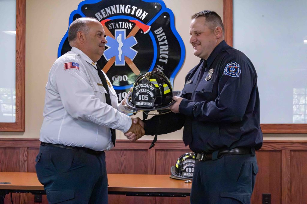 A firefighter in uniform receives a helmet from a man in a uniform with an American flag on his shirt during a ceremony, with a Bennington Station logo in the background.