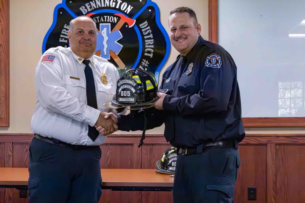 A fire chief and a firefighter are shaking hands while the firefighter holds a fire helmet. The background features a fire station emblem.