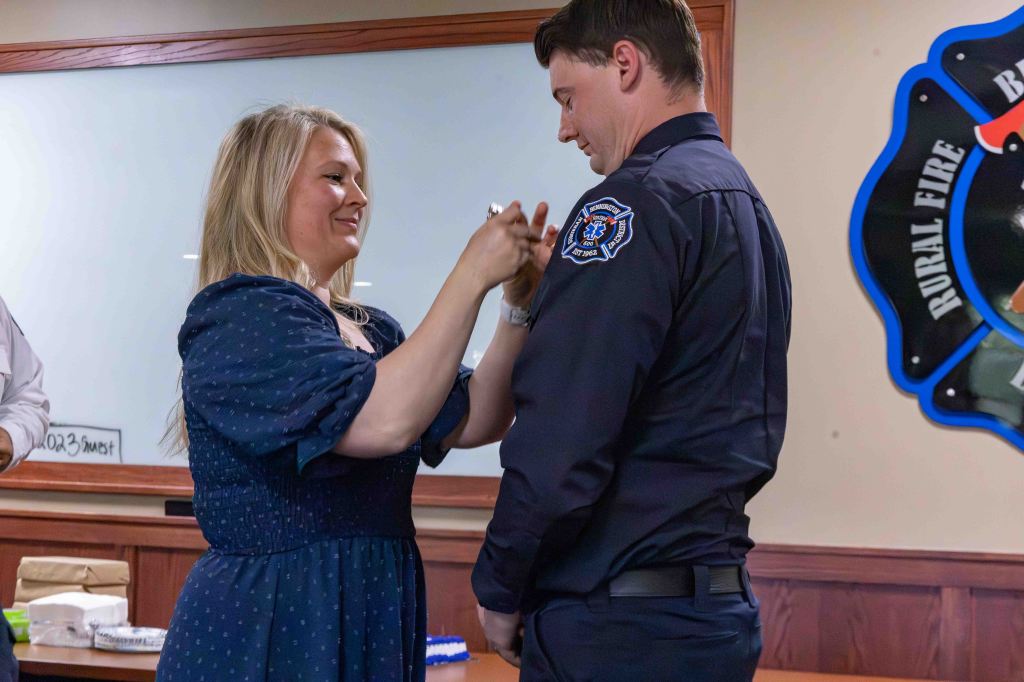 A woman pins a badge on a man in a uniform during a ceremonial event, smiling at each other in a decorated room.