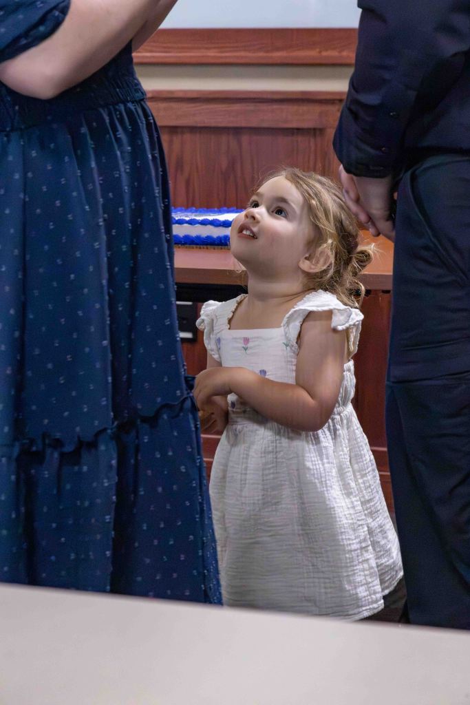 A young girl in a white dress looks up with curiosity at adults nearby, in a formal setting.