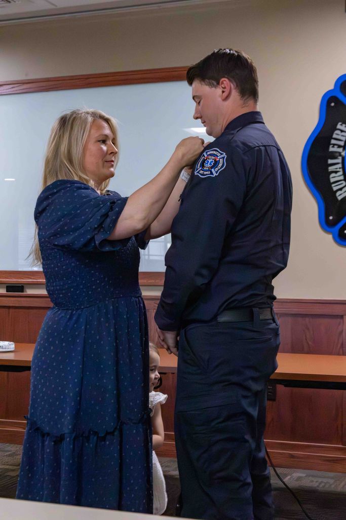 A woman pins a badge onto a man in a uniform during a ceremony, with a young girl peeking out from behind them.