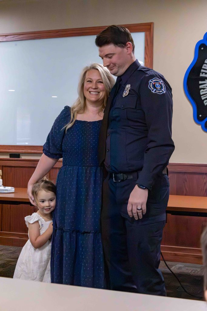 A man in a police uniform stands next to a smiling woman and a young girl, all three posing together in a room with wooden accents.
