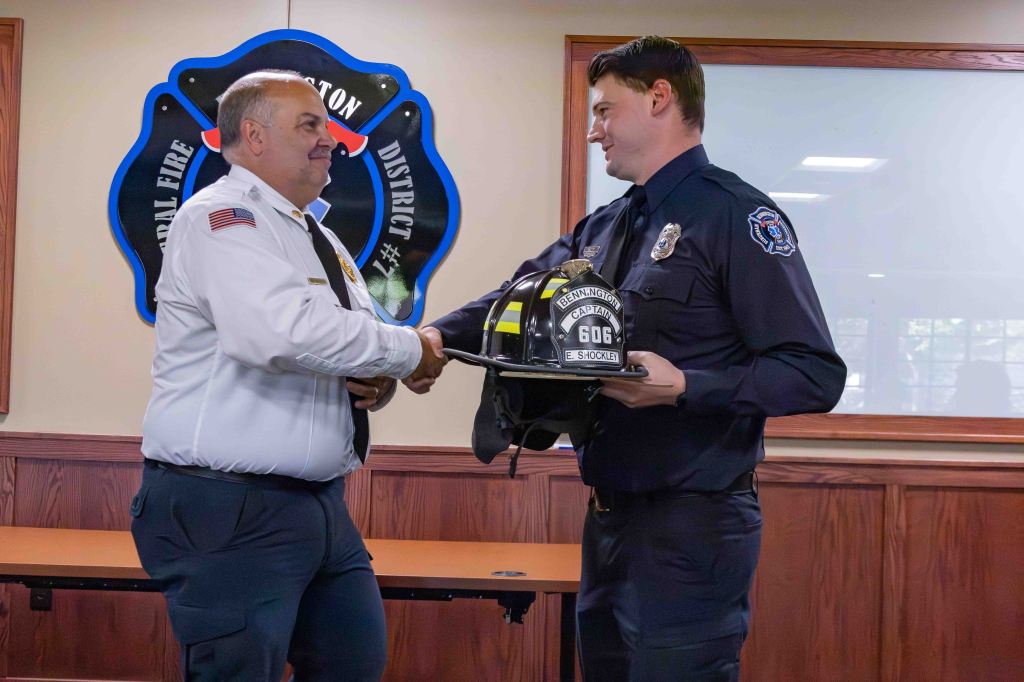 A fire chief shakes hands with a firefighter while presenting him with a helmet during a formal ceremony.