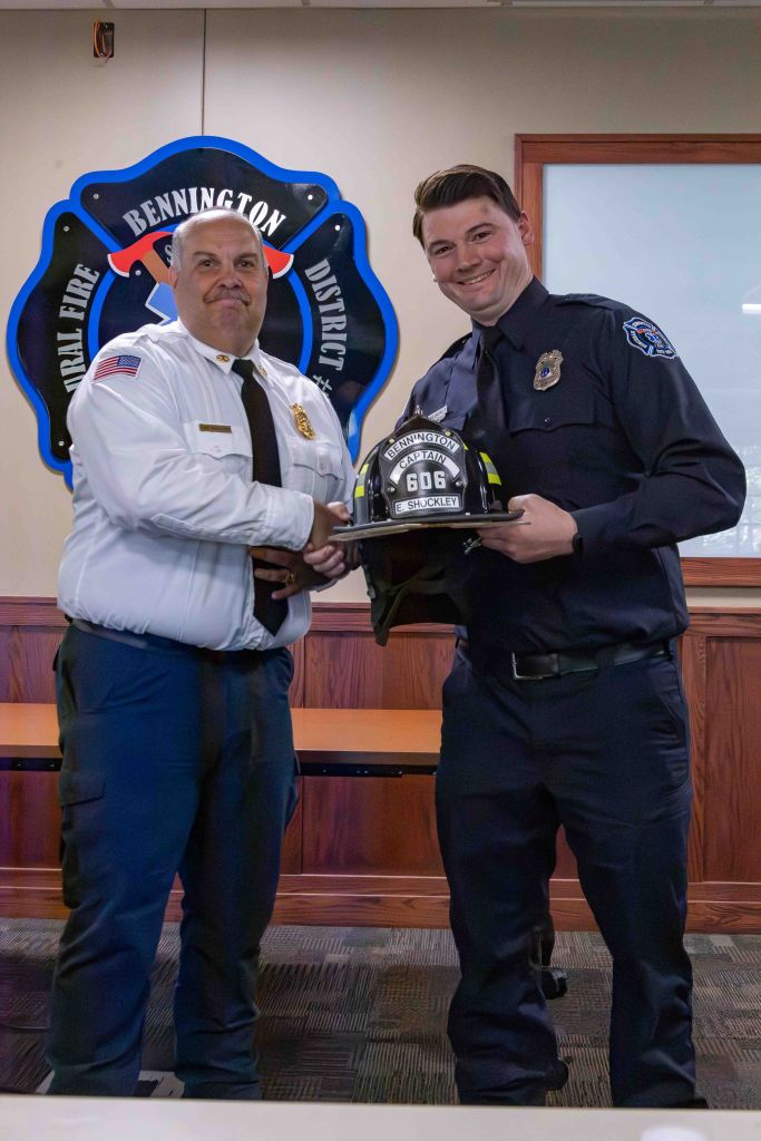 Two firefighters are smiling as one presents a helmet to the other during a ceremony, with a fire department emblem in the background.