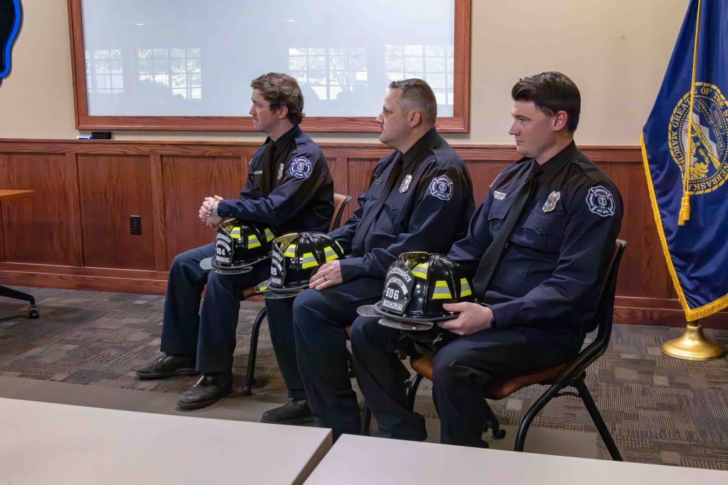 Three fire department personnel sitting in a meeting room, wearing uniforms and holding their helmets.