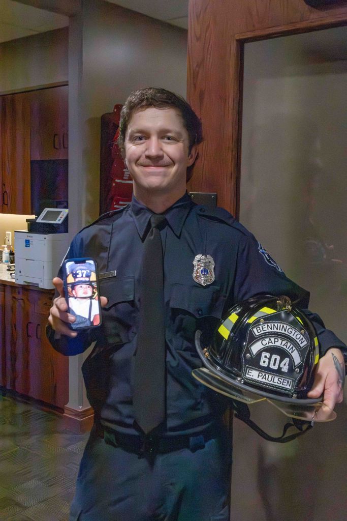 Captain E. Paulsen, a firefighter in uniform, smiles while holding a phone displaying a photo and a firefighter helmet with the name 'Bennington' and designation 'Captain 604'.