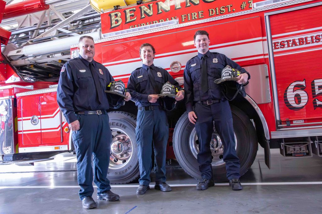 Three firefighters standing in front of a red fire truck, each holding a helmet. They are dressed in navy blue uniforms with badges.