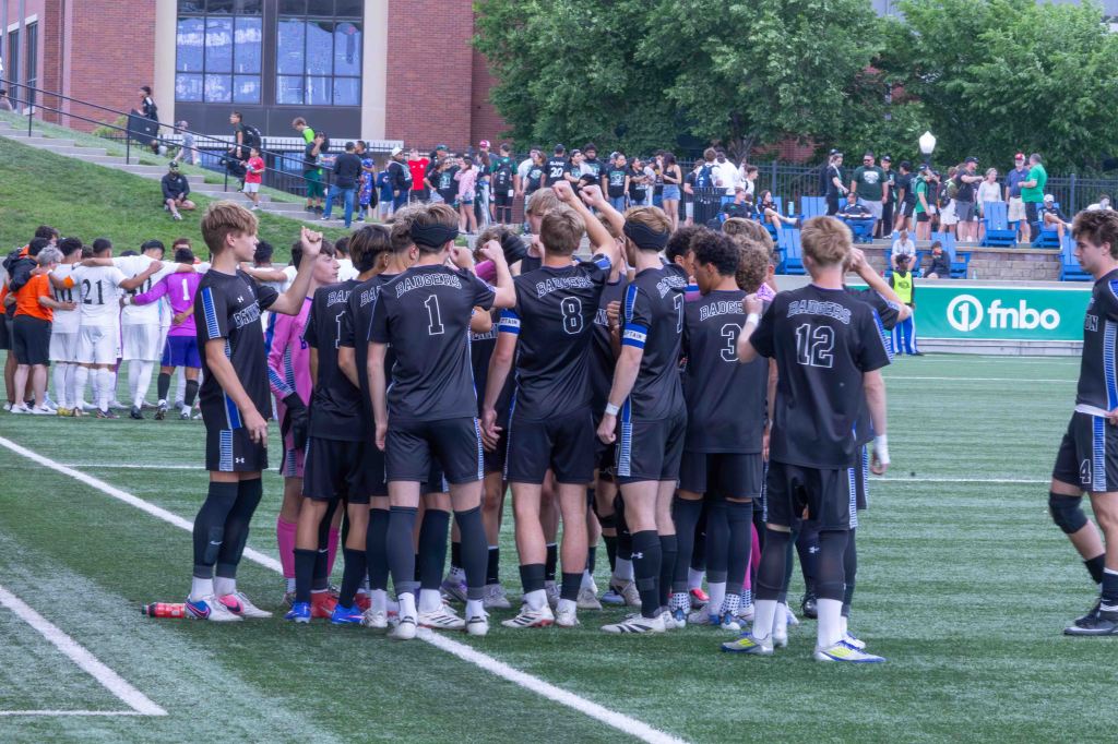 A soccer team in black uniforms huddles together on the field, preparing for a game, while another team in white uniforms stands in the background.