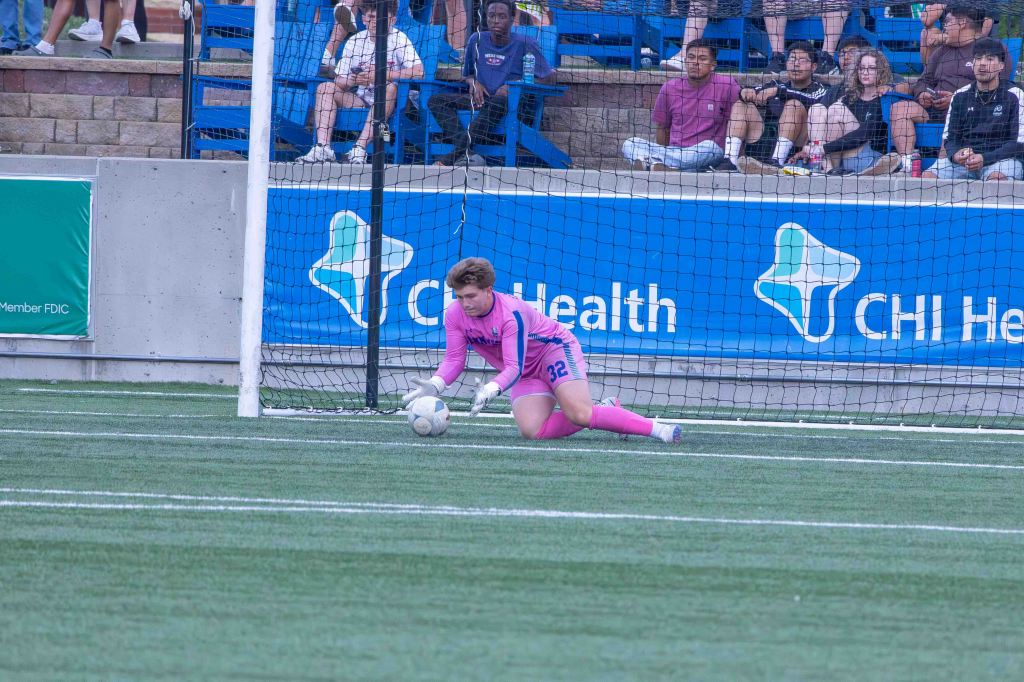 A female soccer goalkeeper in a pink uniform is diving to catch the ball during a game, with spectators seated in the background.