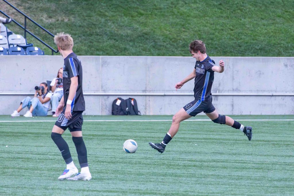 A soccer player in a black uniform kicks a soccer ball on a green field while another player is in the background, and a few spectators are seated on the sidelines.
