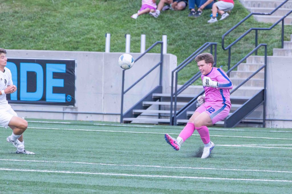 A soccer goalkeeper in a pink uniform is about to kick a ball away, while an opposing player in white is positioned to the side. In the background, spectators are watching the game on a grassy hillside.