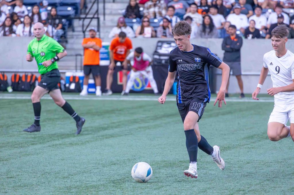 A youth soccer player in a black jersey dribbles a soccer ball on a grassy field during a match, with a referee in a green uniform nearby and spectators in the background.