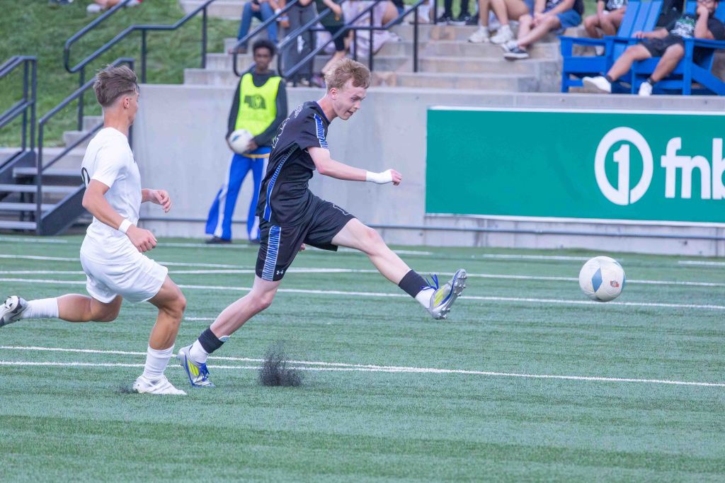A young soccer player in a black uniform kicking a soccer ball on a field, while another player in a white uniform runs nearby.