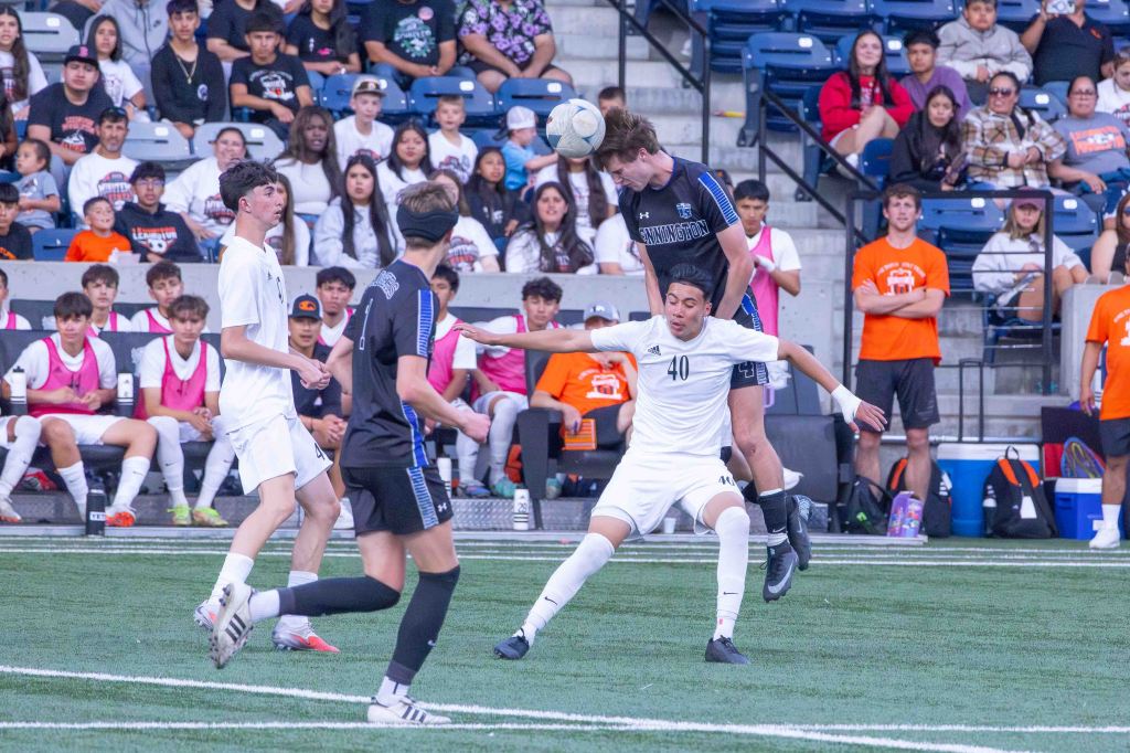A soccer match in progress, featuring players in black and white uniforms competing for the ball. One player is jumping to head the ball while another is positioned on the ground. The stands are filled with spectators watching the game.