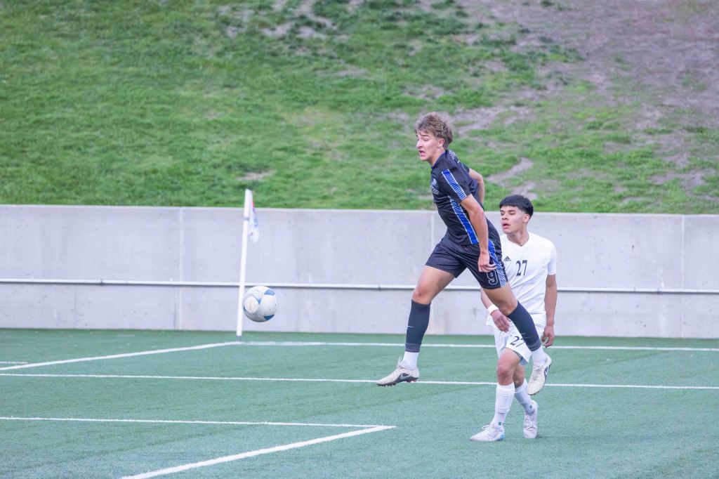 A soccer player in a blue uniform jumps to head the ball while an opponent in a white uniform attempts to defend.
