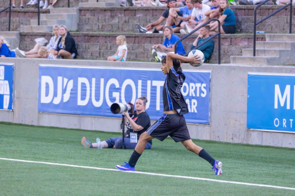 A young male soccer player performing a throw-in during a match, with fans seated on bleachers in the background.