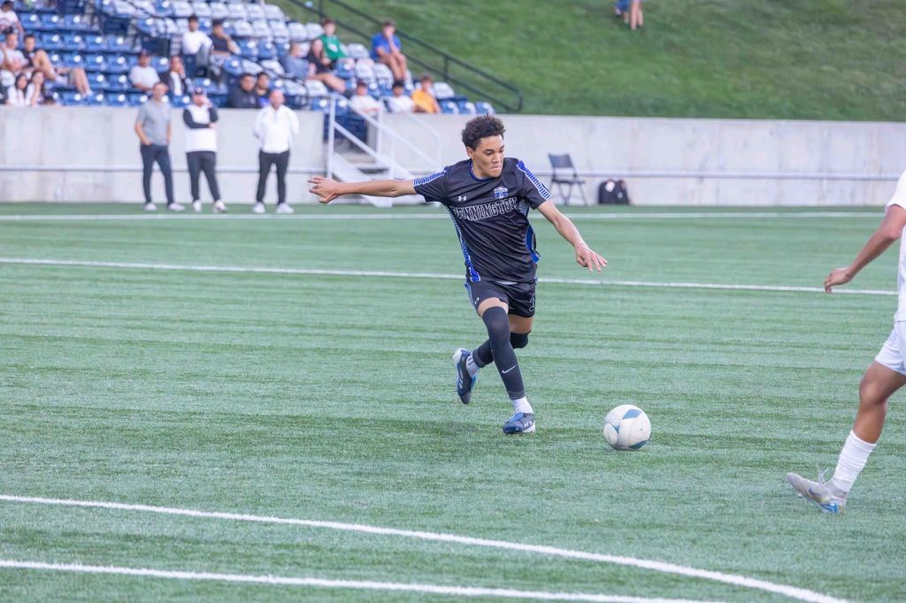 A soccer player in a black uniform is kicking a soccer ball on a green field during a match, with spectators in the background.