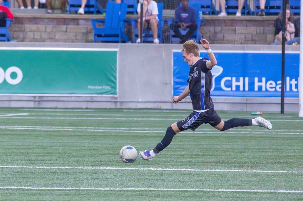 A soccer player in a black jersey executes a kick on the field with an outstretched leg, while spectators watch in the background.