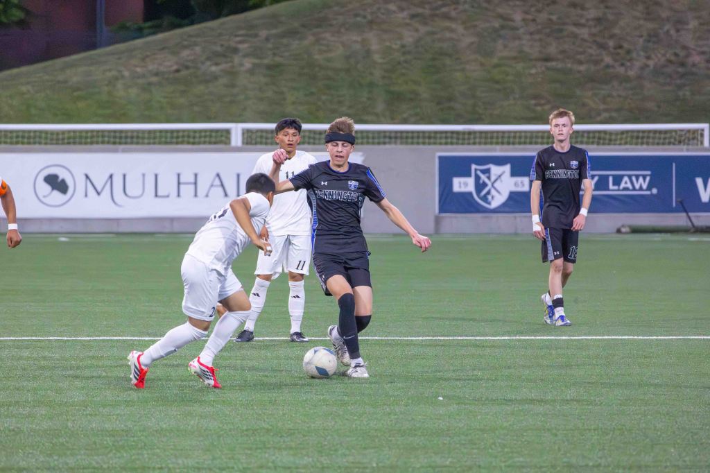 A soccer match in action, featuring players in black and white uniforms. One player in black is dribbling the ball while another in white attempts to steal it. In the background, other players are positioned ready for play.