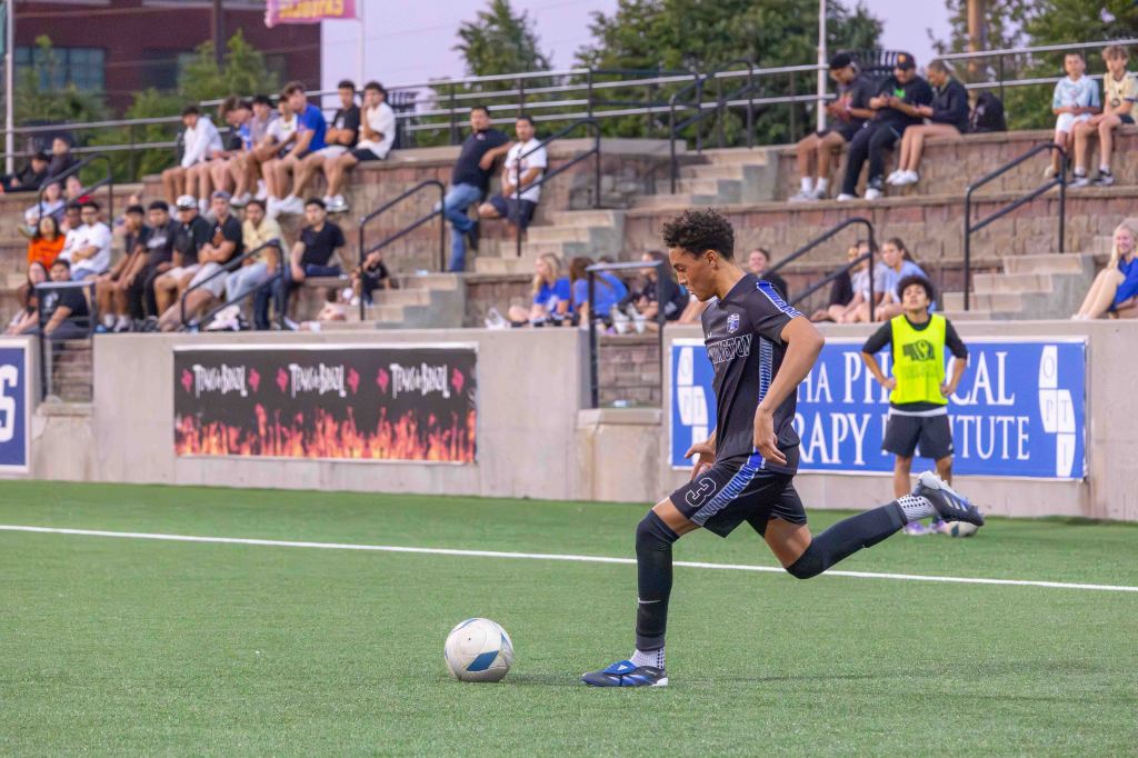 A soccer player in a black uniform prepares to kick a ball on a field, with spectators in the background watching the game.