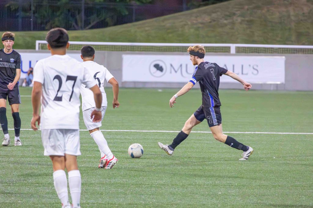 A soccer player in a black uniform with the number 7 kicks a ball on a green field, while two opposing players in white uniforms, one with the number 27, look on.